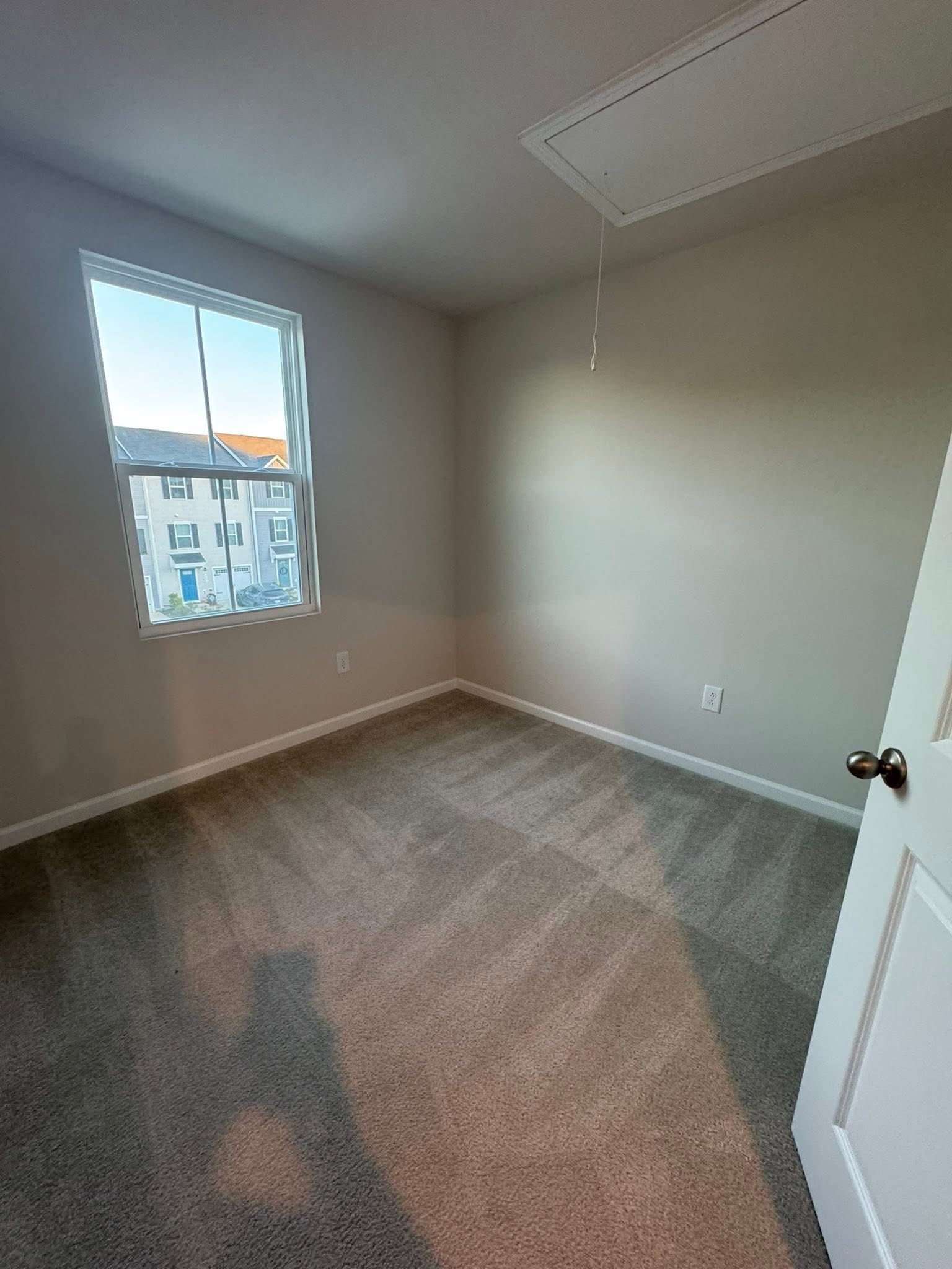 An empty bedroom with neutral walls, grey carpet, a single window overlooking nearby buildings, and an attic access panel.