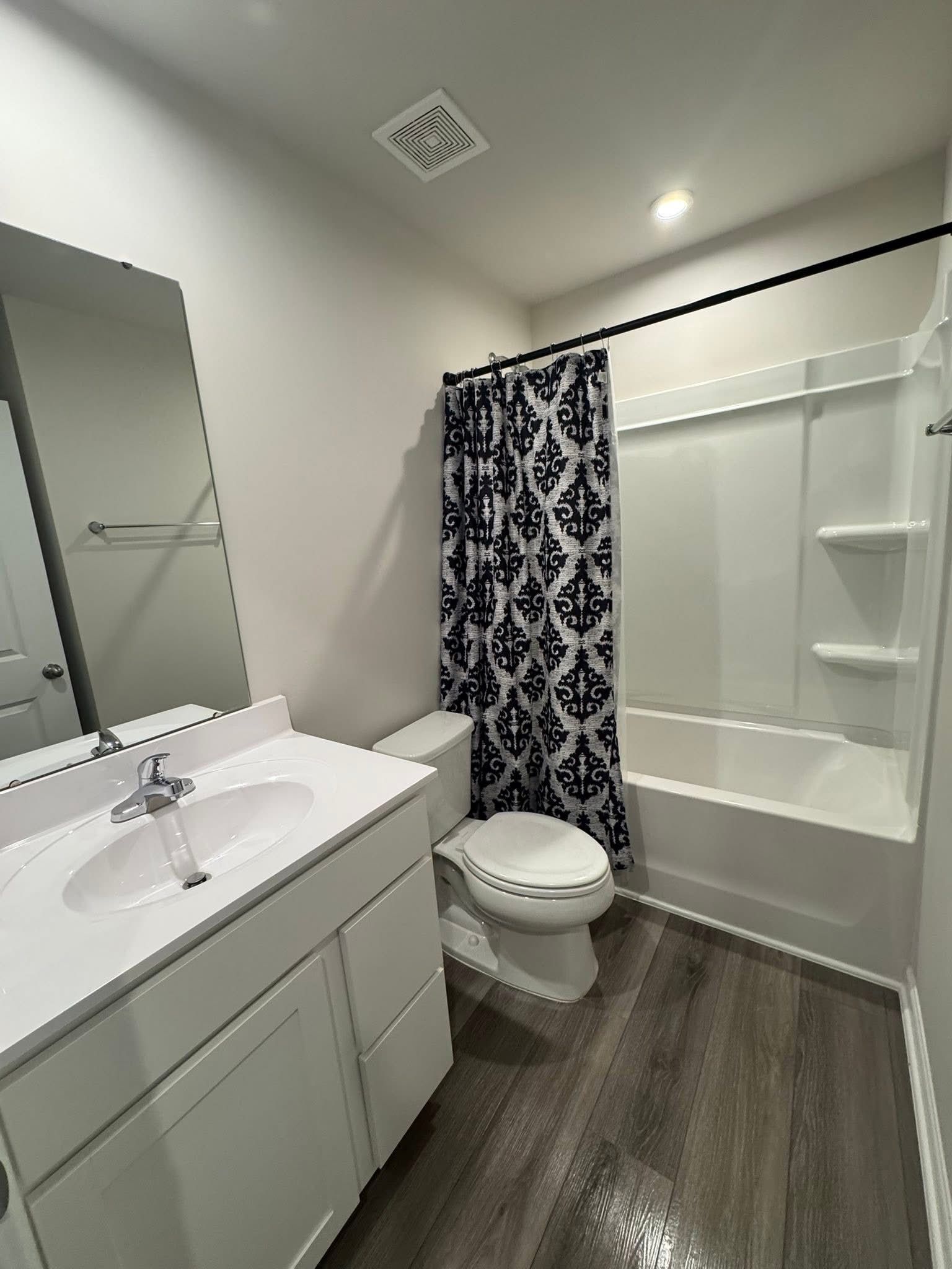 A modern bathroom featuring a white vanity with a chrome faucet, a patterned shower curtain, and wood-look flooring.