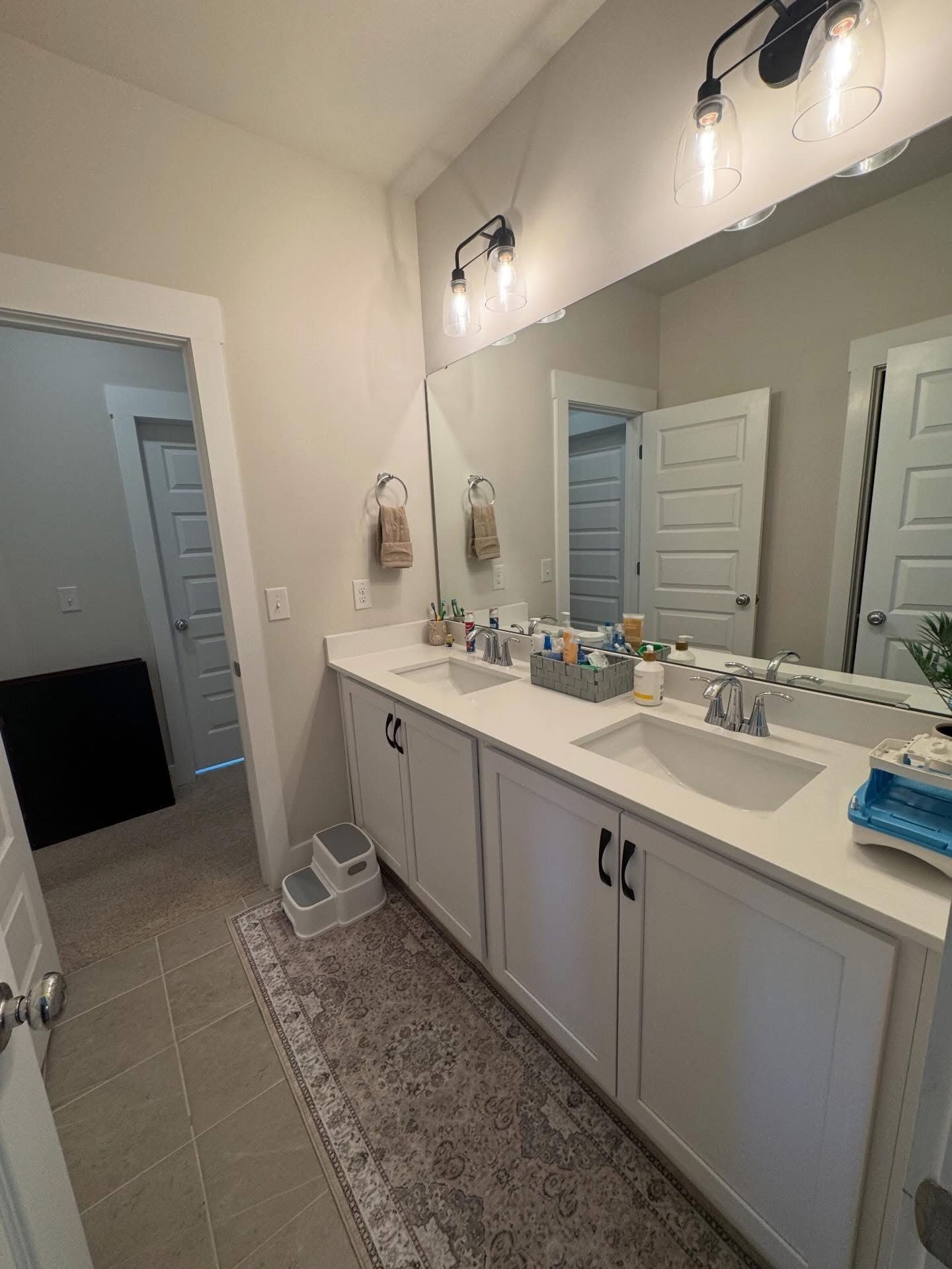 A bright, modern bathroom featuring a white double-sink vanity, a large wall mirror, and an adjacent open doorway.