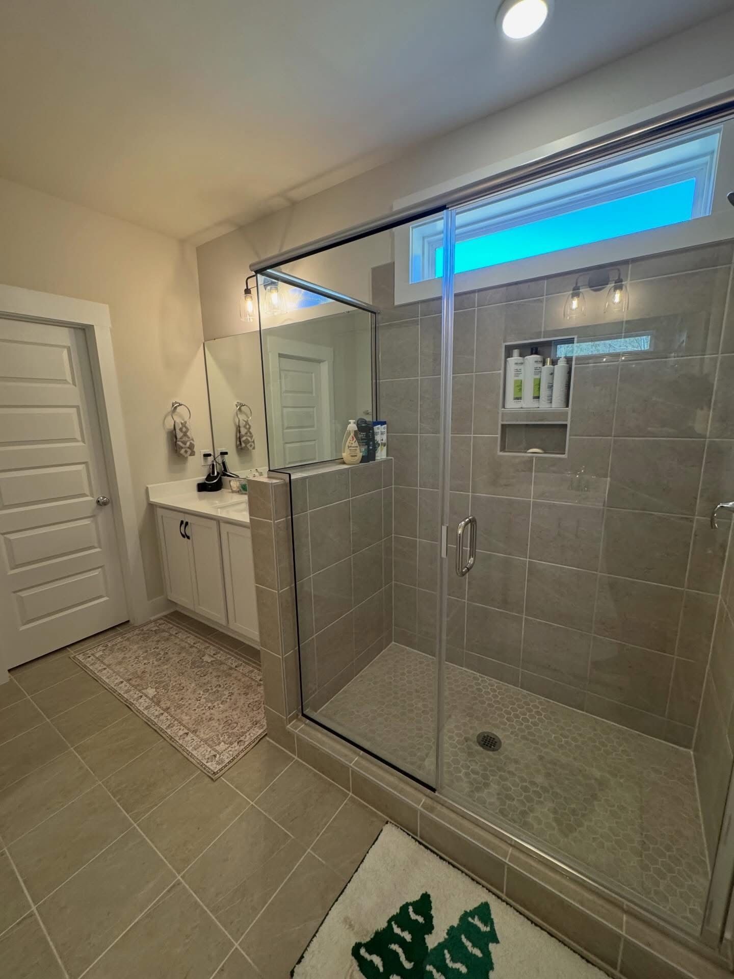 Modern bathroom with a tiled walk-in glass shower, a white vanity cabinet, and a door on the left.