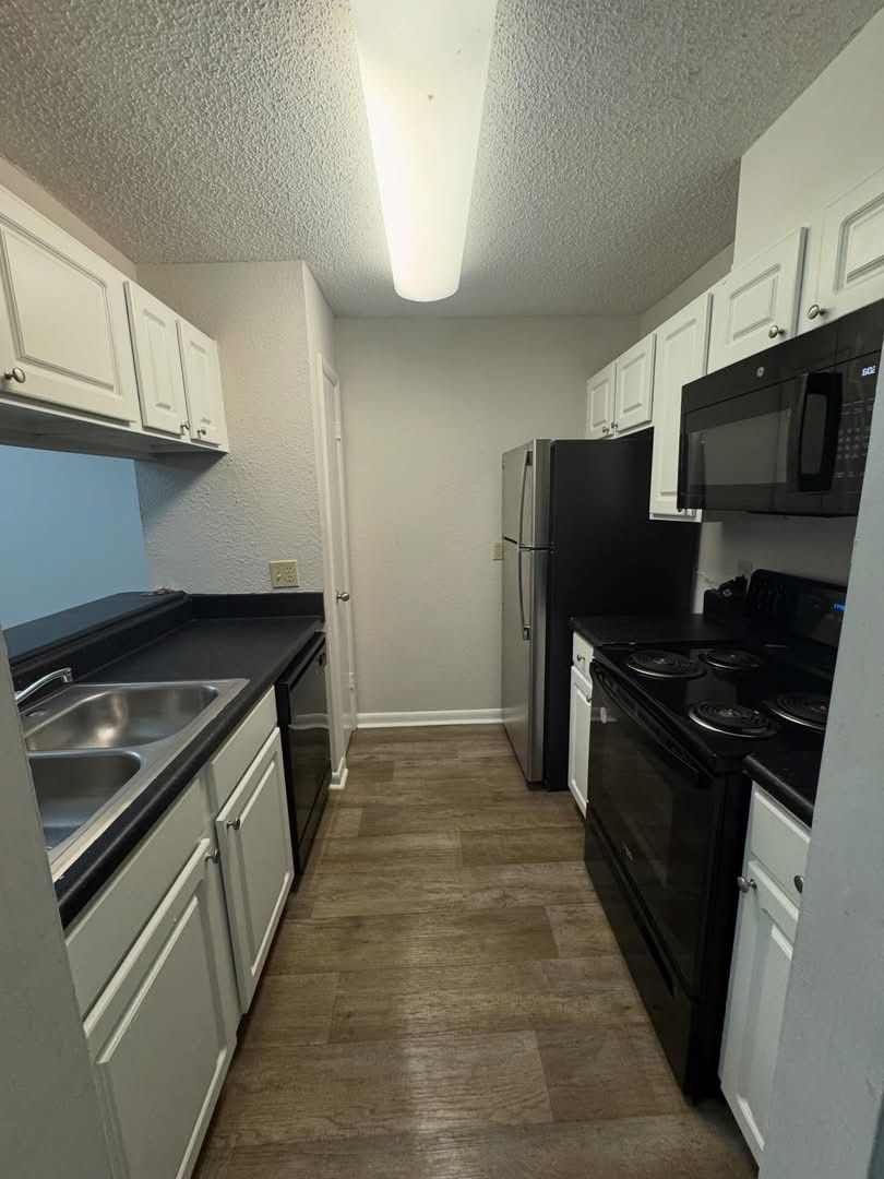 A galley kitchen with white cabinets, dark countertops, a double sink, black appliances, and light wood-look flooring.