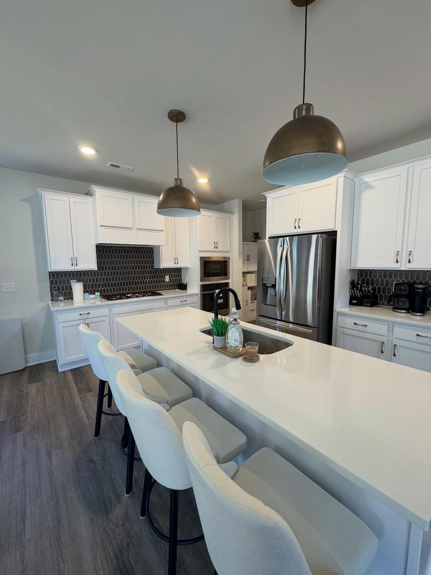 A modern kitchen featuring white cabinets, a long white countertop island with four bar stools, and two pendant lights.