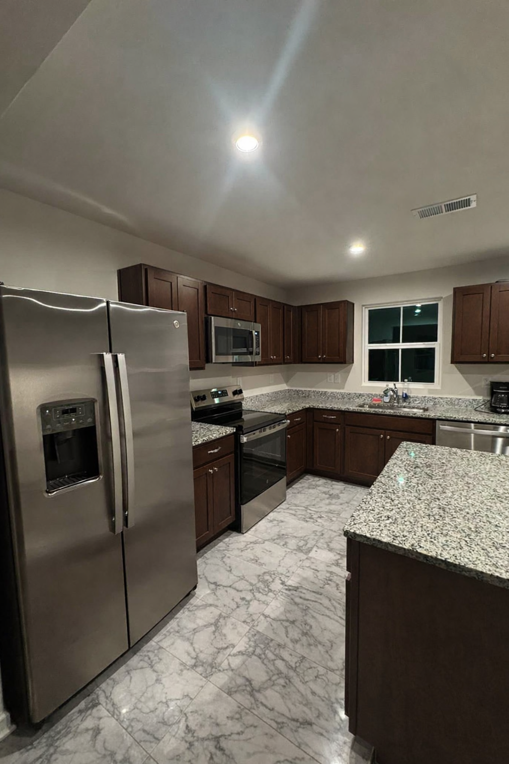 Modern kitchen with dark wood cabinets, granite countertops, stainless steel appliances, and gray marbled tile flooring.