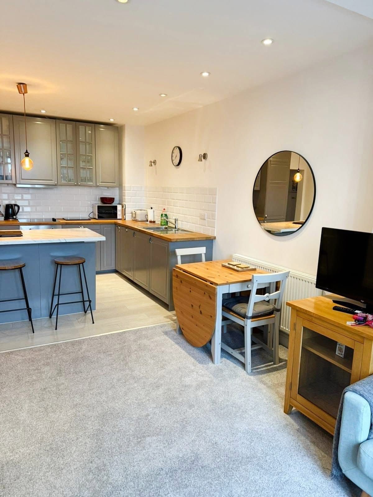 A modern open-plan kitchen and dining area with a blue island, wooden table, round wall mirror, and a television on a unit.