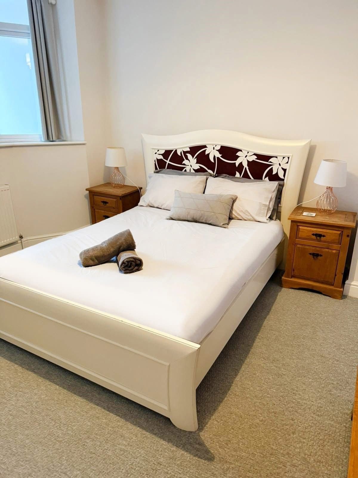 A white bed with a floral headboard and two rolled towels sits between two wooden nightstands in a carpeted room.