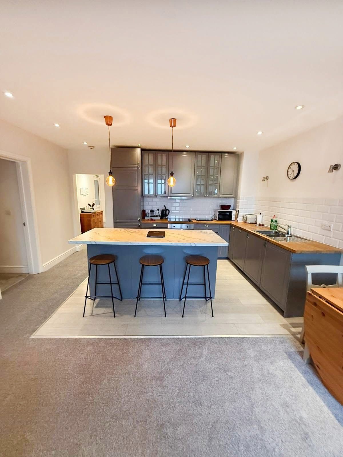 A kitchen with a large gray island, three stools, and wooden countertops, situated in an open-plan room with carpet.