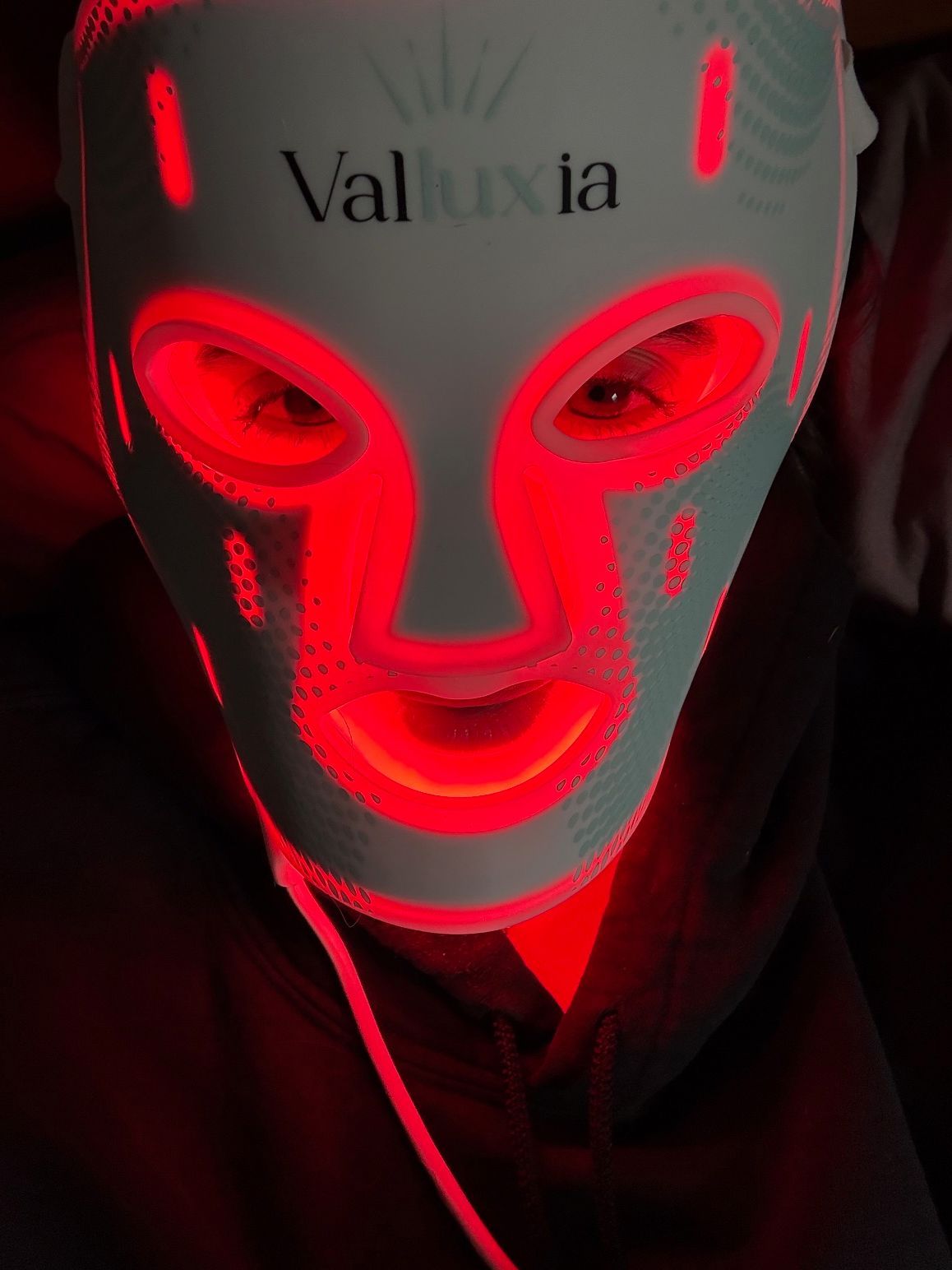 Person wearing a white LED light therapy mask emitting red light. The mask covers the face.