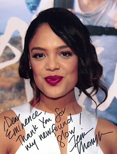 Tessa Thompson with dark hair, dark pink lipstick, smiling, holding an autograph.