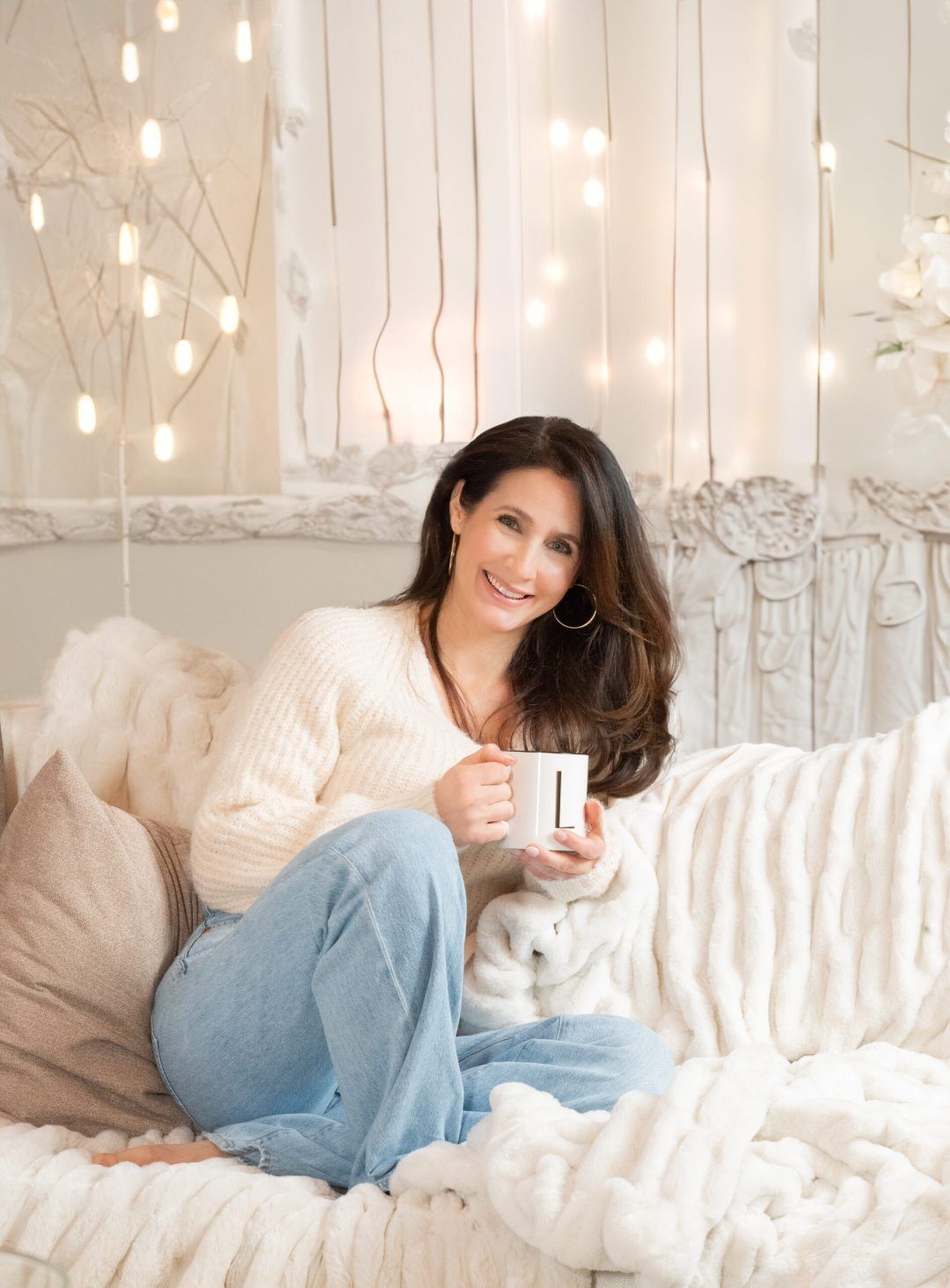 Woman in a cream sweater and jeans smiles, holding a mug, sitting on a bed with cozy blankets, string lights in background.
