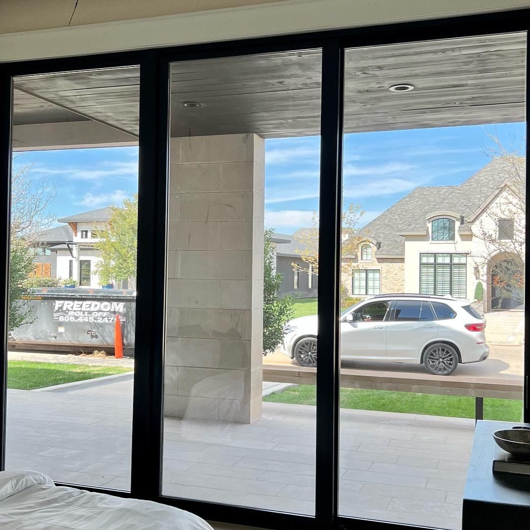 Interior view through black-framed sliding glass doors to a sunny suburban street and parked white car