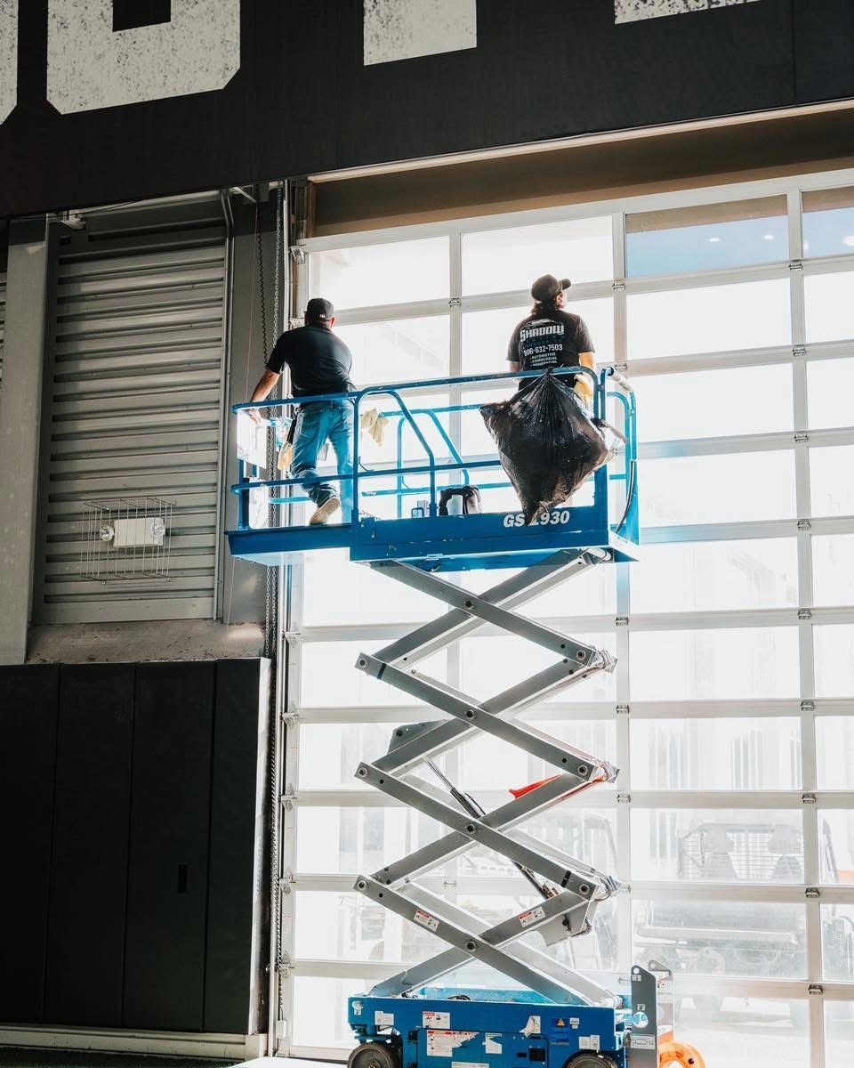 Two workers on a blue scissor lift inside a building, working near a large garage door.