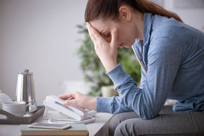 Woman at a desk, holding her forehead while looking at a phone and papers, appearing stressed.