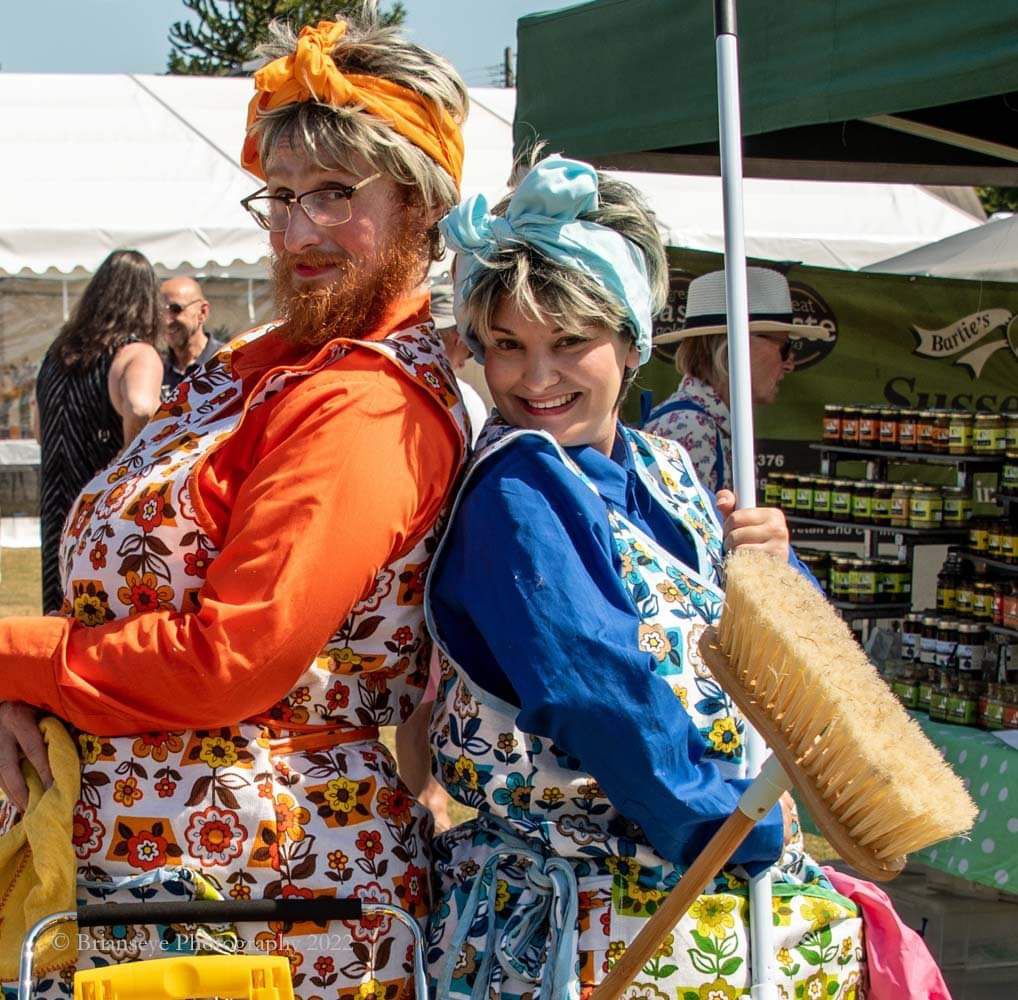Two performers wearing 1950's aprons stand back to back smiling. One is holding a broom