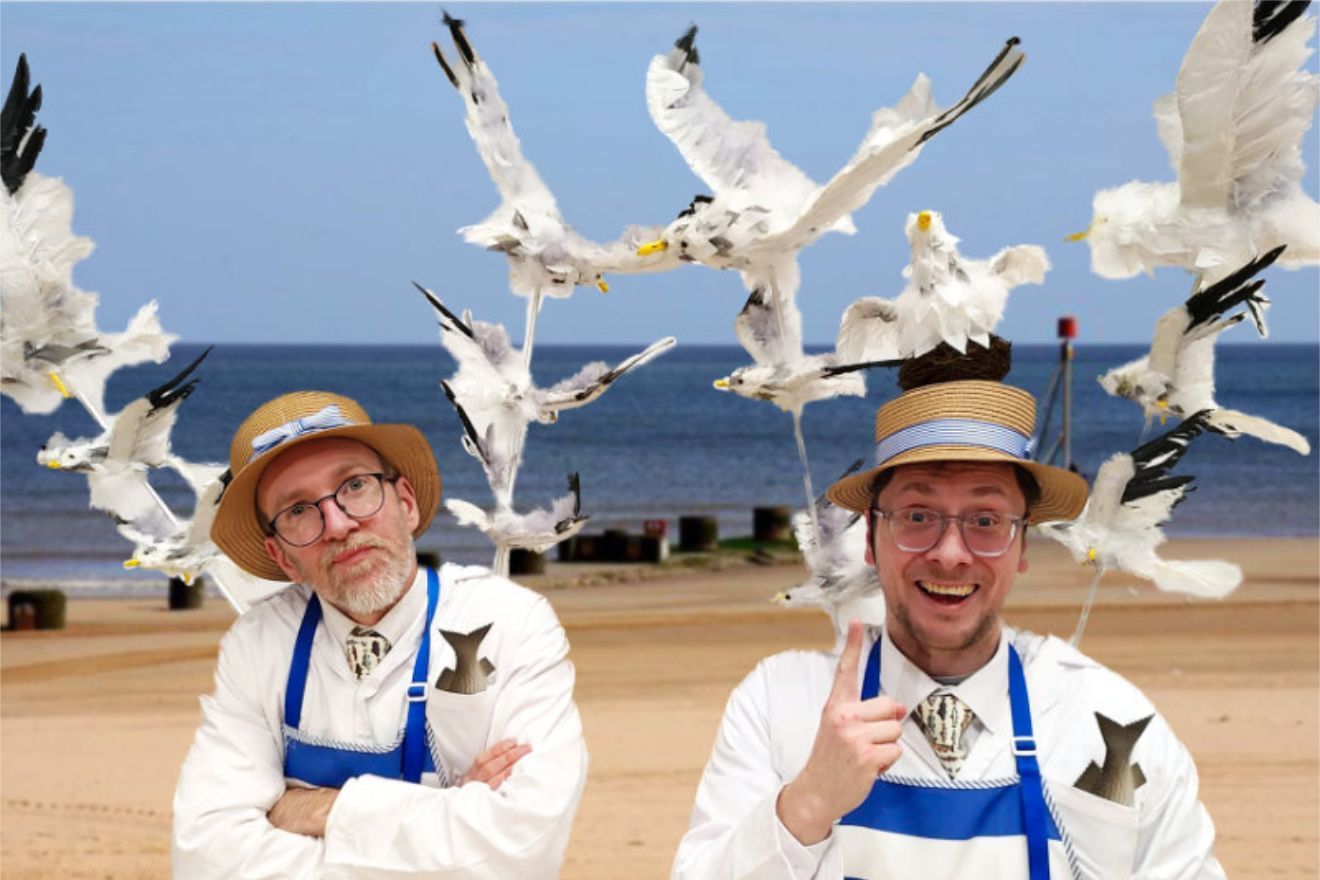 Two performers dressed as fishmongers. One is unhappy and has his arms crossed. the other looks pleased and is merrily pointing at the seagulls above