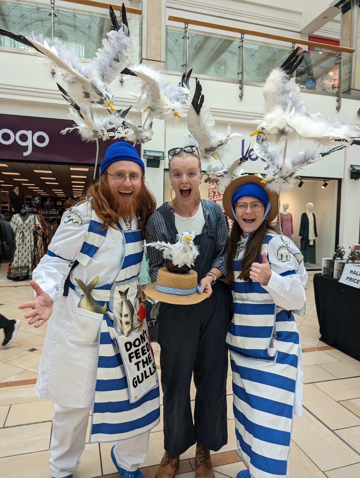 Two performers dressed as fishmongers, wearing backback with seagulls raised above their heads, stand with a smiling member of the public