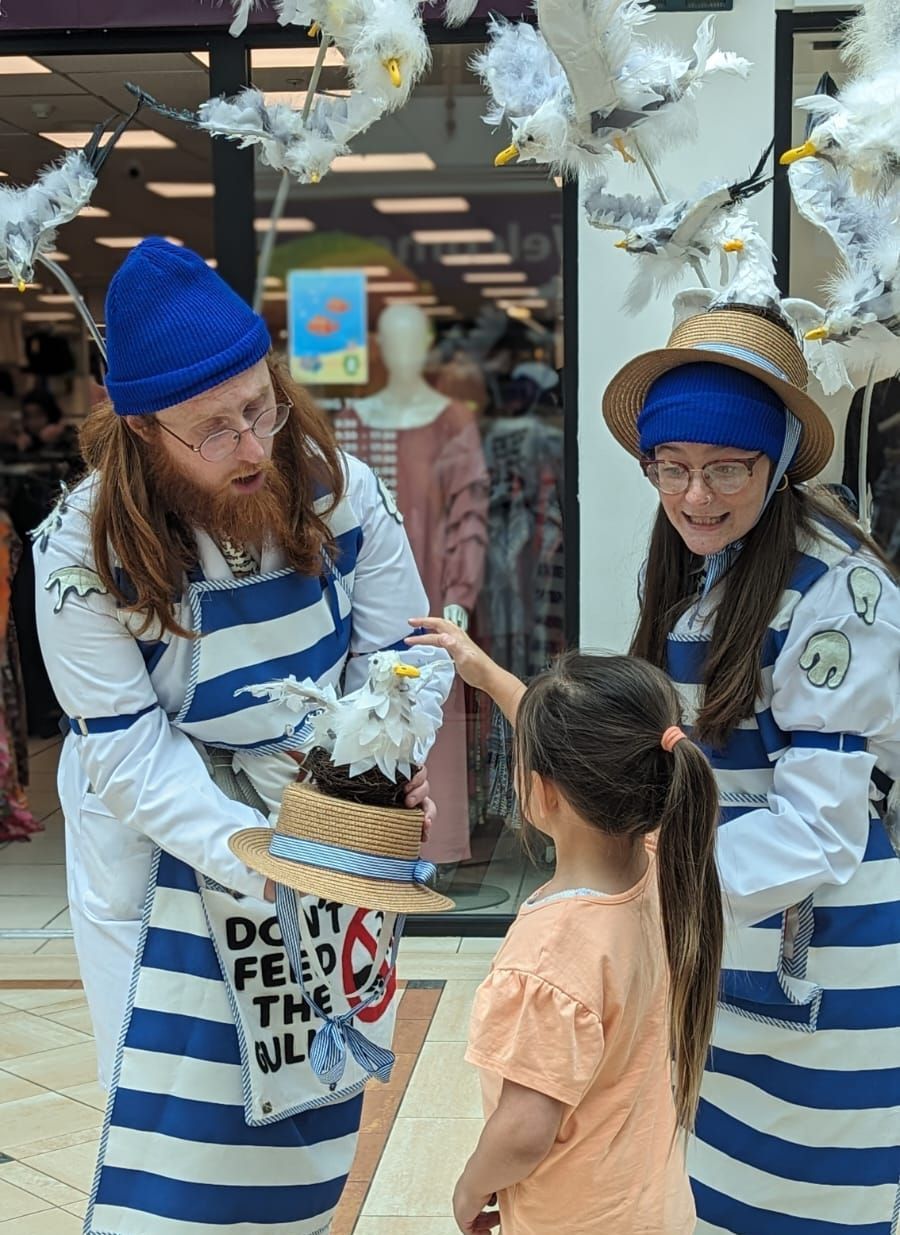 Two performers dressed as fishmongers, wearing backback with seagulls raised above their heads, show a seagull puppet to a young child