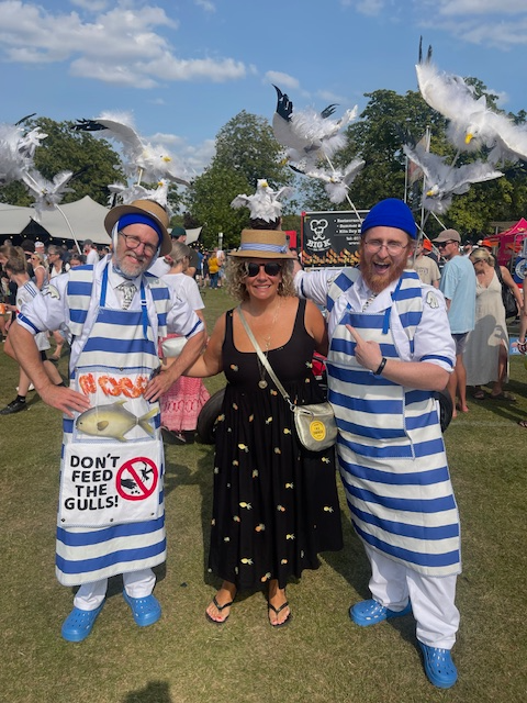 Two performers dressed as fishmongers, wearing backback with seagulls raised above their heads, stand with a smiling member of the public wearing a straw boater hat with a nesting seagul on it