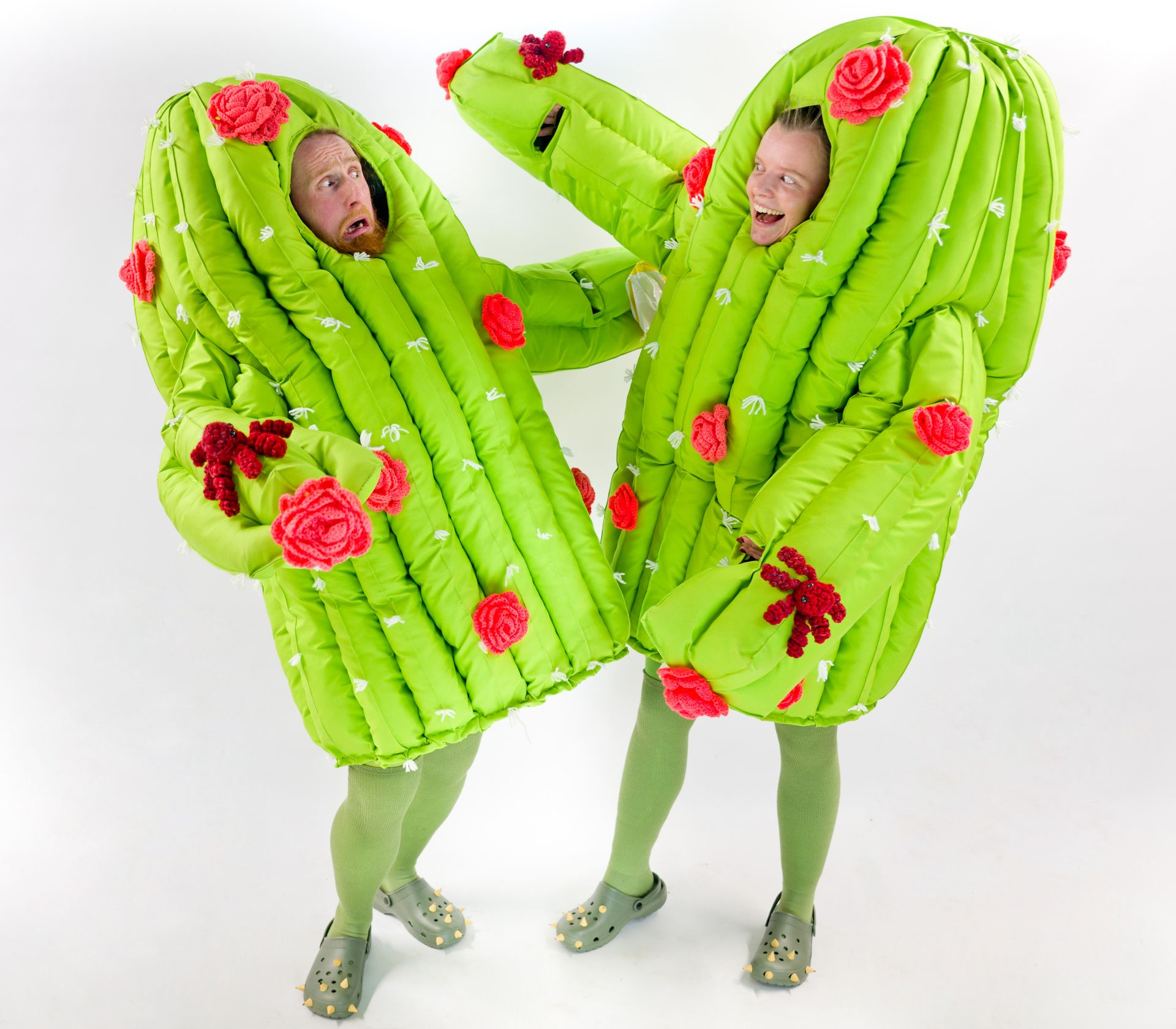 Two performers dressed as oversized green cactus characters with pink flowers strike a playful scene, one cacti is joyously going in for a hug while the other looks concerned