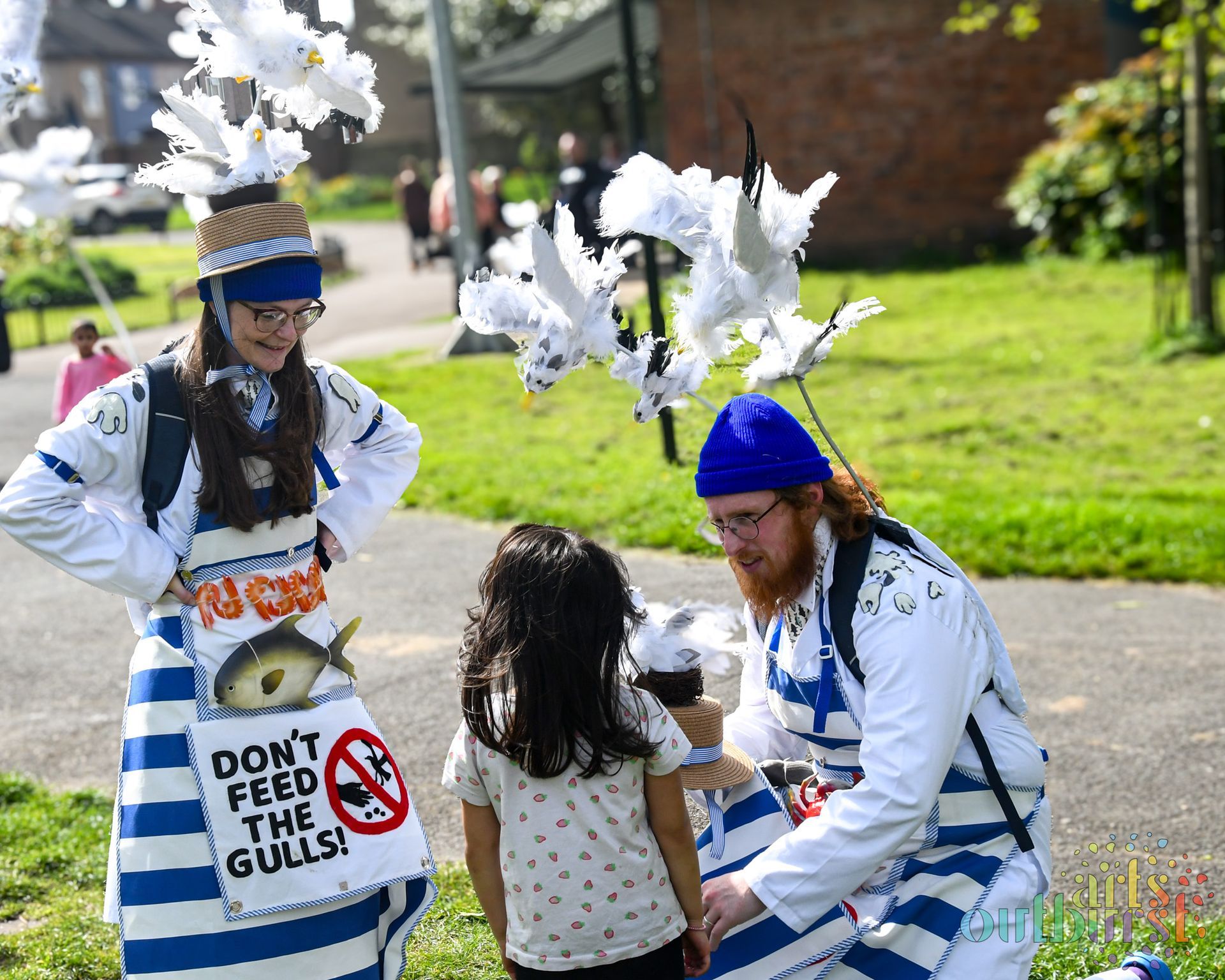 Two performers dressed as fishmongers, wearing backback with seagulls raised above their heads, show a seagull puppet to a young child