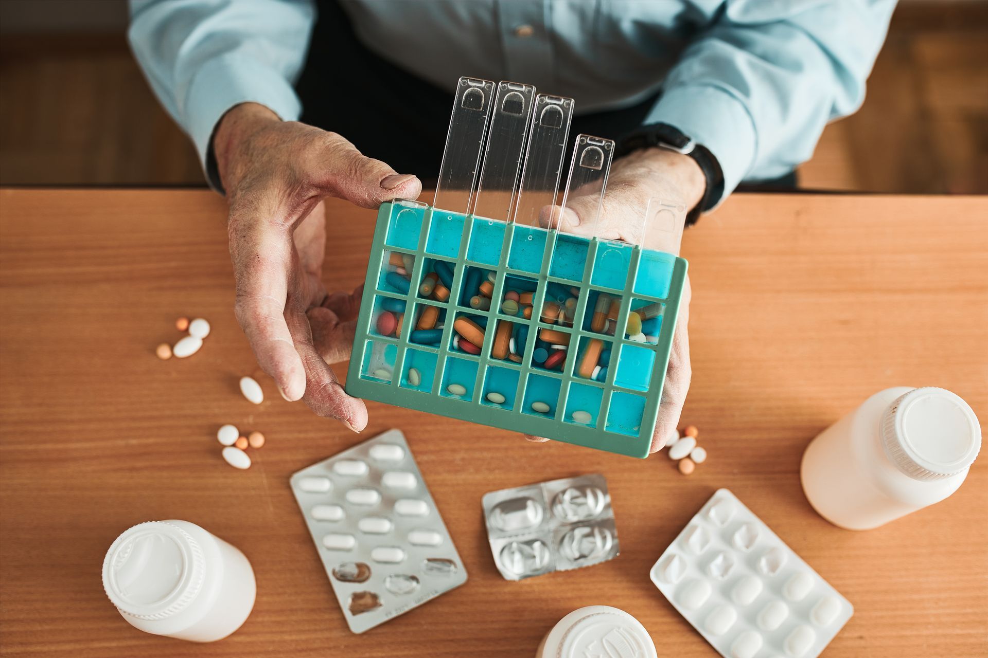 Hands pouring pills into a turquoise weekly pill organizer on a wooden table with scattered pill bottles and tablets