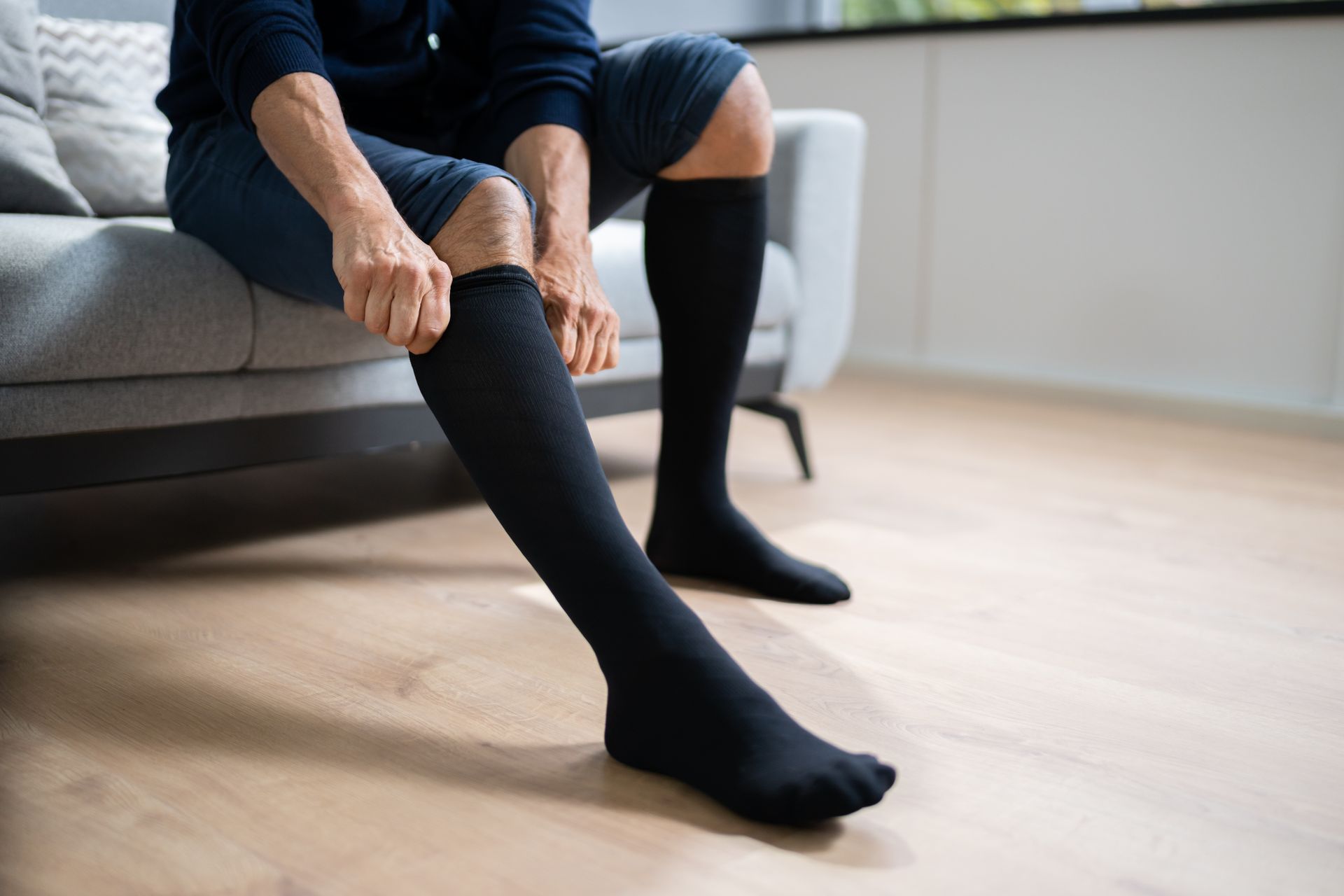 Person sitting on a gray sofa pulling on black compression socks in a bright living room