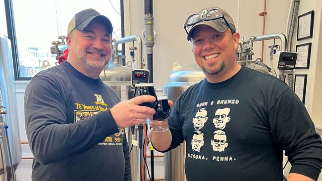 Two men in a brewery toast with glasses of dark beer. Both are smiling, wearing hats and casual shirts.