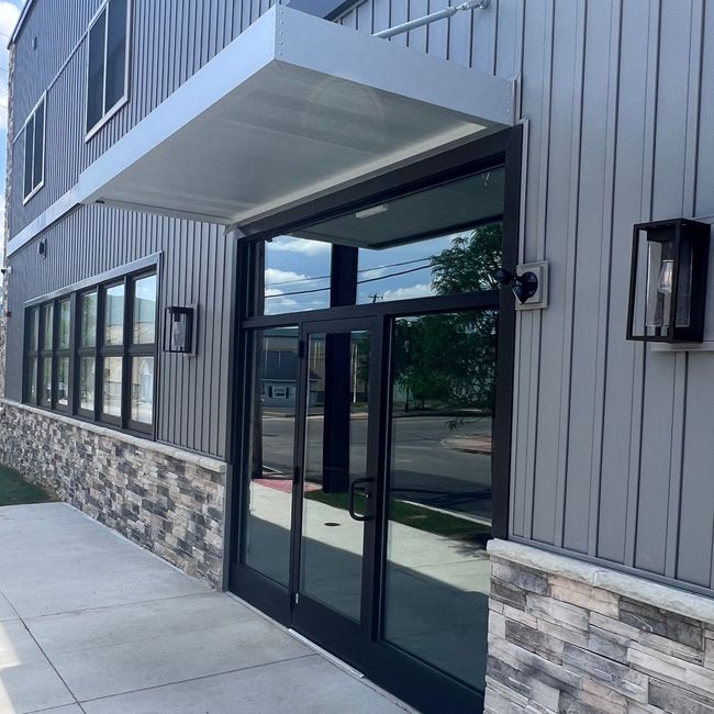 Building entrance with glass doors, awning, and sconces. Gray and stone exterior.