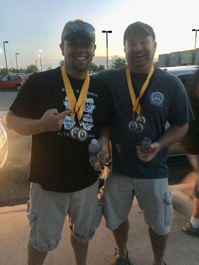 Two men wearing medals, smiling outdoors. One points to his medal; wearing shorts and t-shirts.
