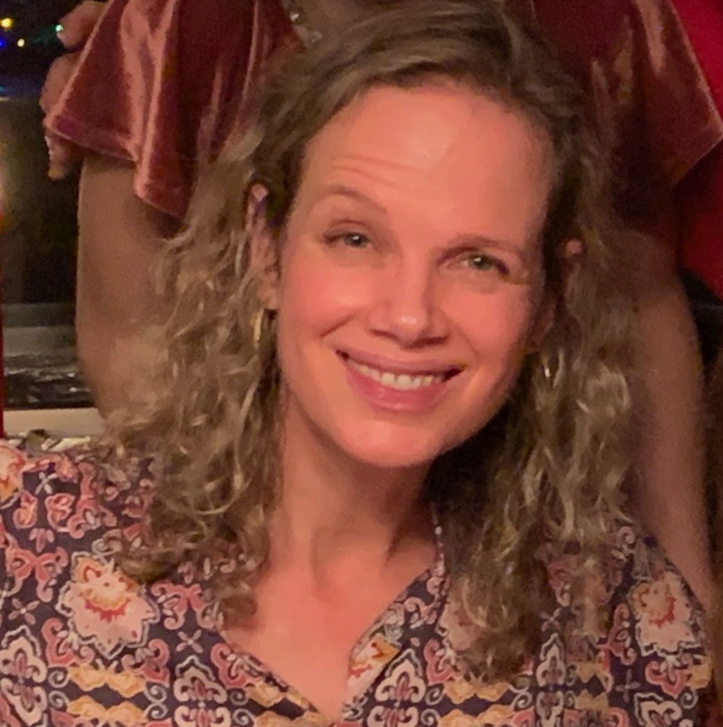 Smiling woman with curly hair wearing a patterned blouse against a plain background