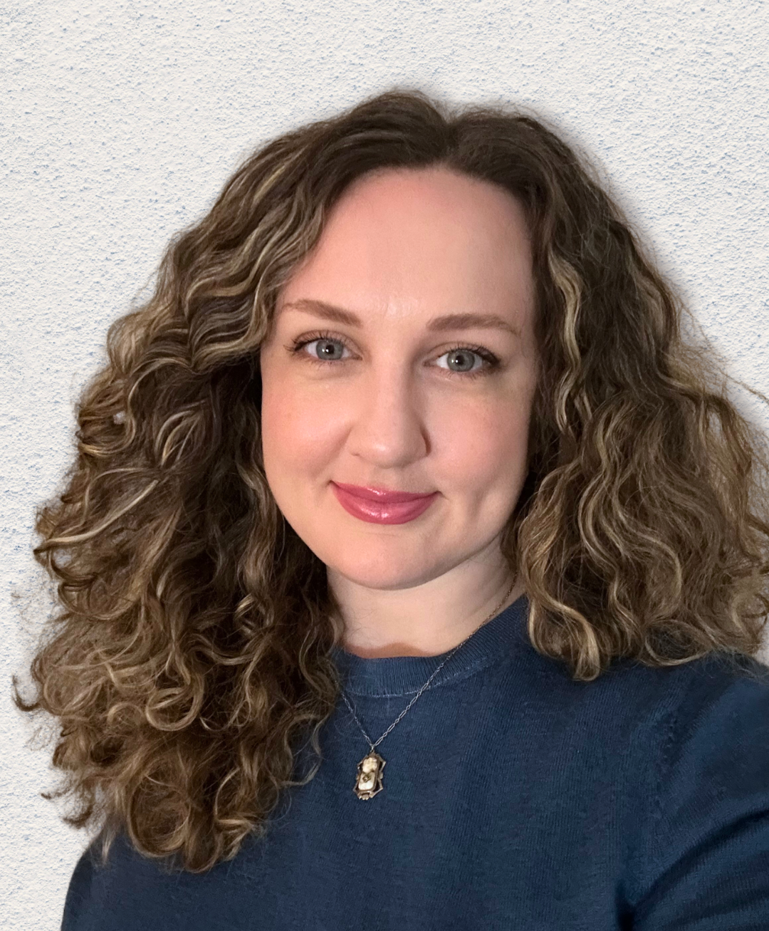 Portrait of a smiling person with curly hair wearing a blue top and pendant necklace against a light wall.
