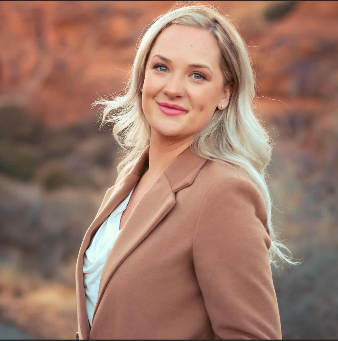 Smiling blonde woman in a tan coat against a blurred rocky outdoor background