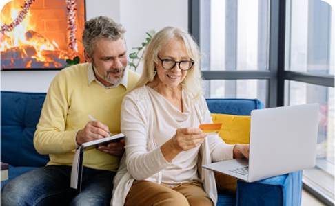 Older couple using a laptop, woman holding credit card, man taking notes, sitting on a blue couch indoors.