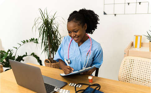 Smiling Black woman in blue scrubs with stethoscope, writing on a clipboard, working on a laptop at a desk.