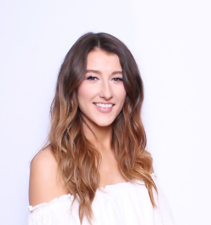 Portrait of a smiling person with long wavy brown hair wearing a white off-shoulder top
