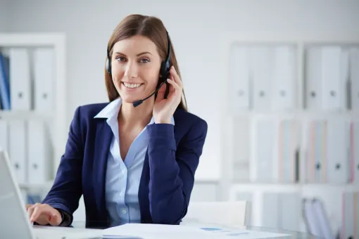 Woman in a navy blazer speaking on a headset at a desk in a bright office