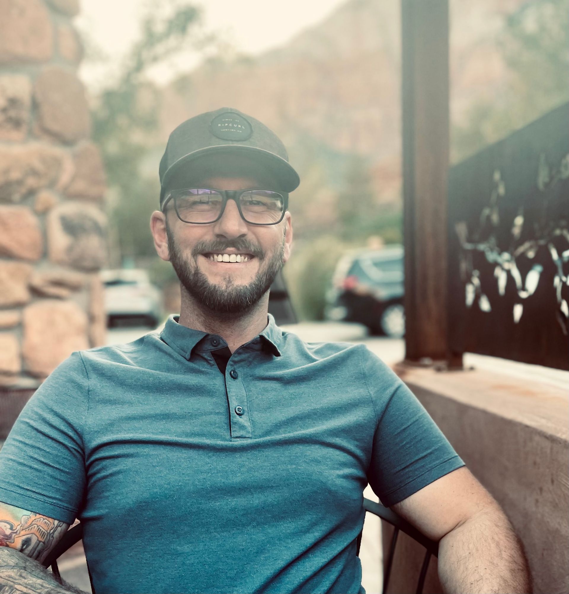 Smiling man in a green cap and glasses, seated outdoors beside a stone wall and black fence.