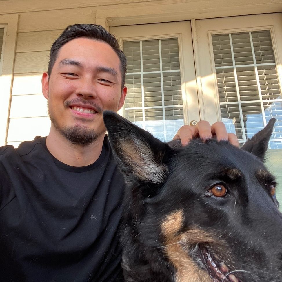 Man smiling beside a black German Shepherd on a porch, hand resting on the dog’s head
