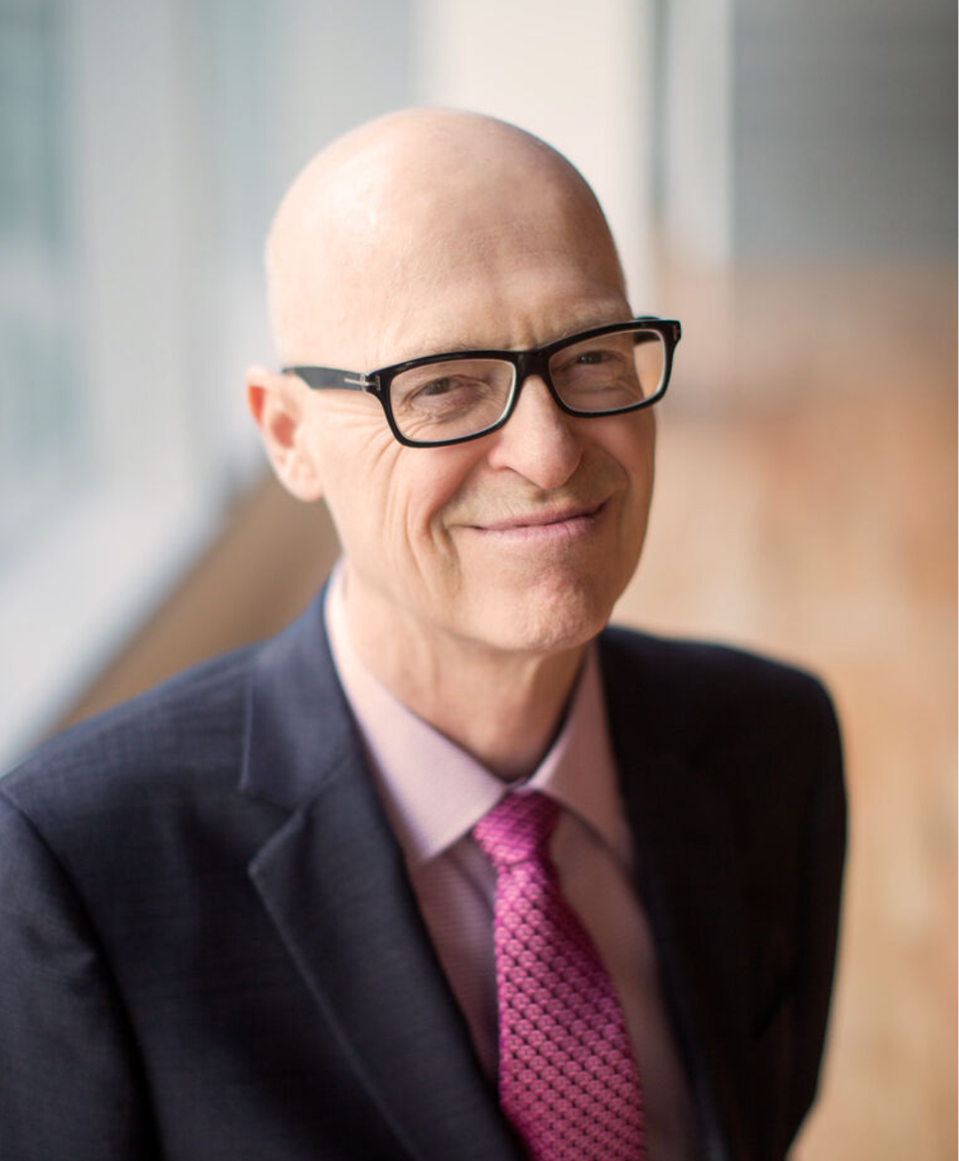 Smiling bald man in glasses wearing a suit and pink tie, standing indoors by a window.