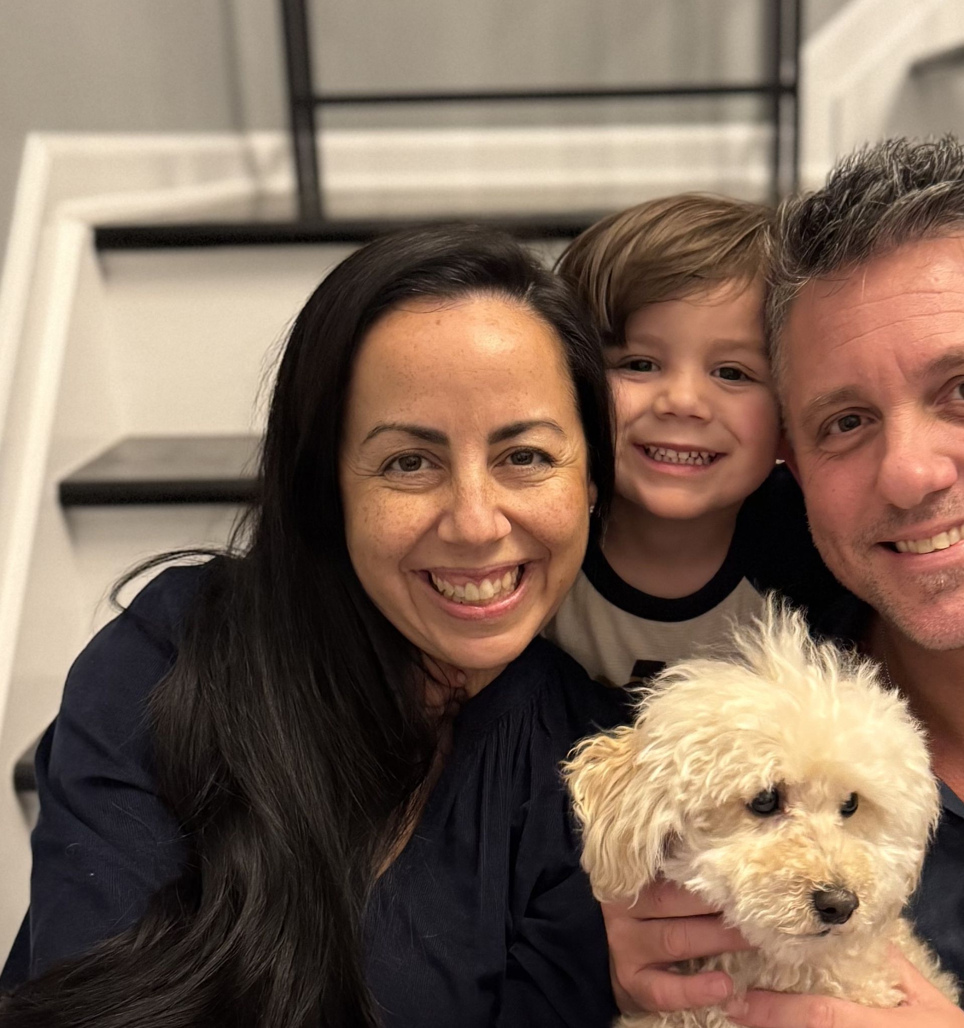 Family selfie with a smiling woman, child, man, and small white dog on stairs