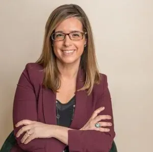 Woman with glasses, wearing a burgundy blazer, smiling with arms crossed.