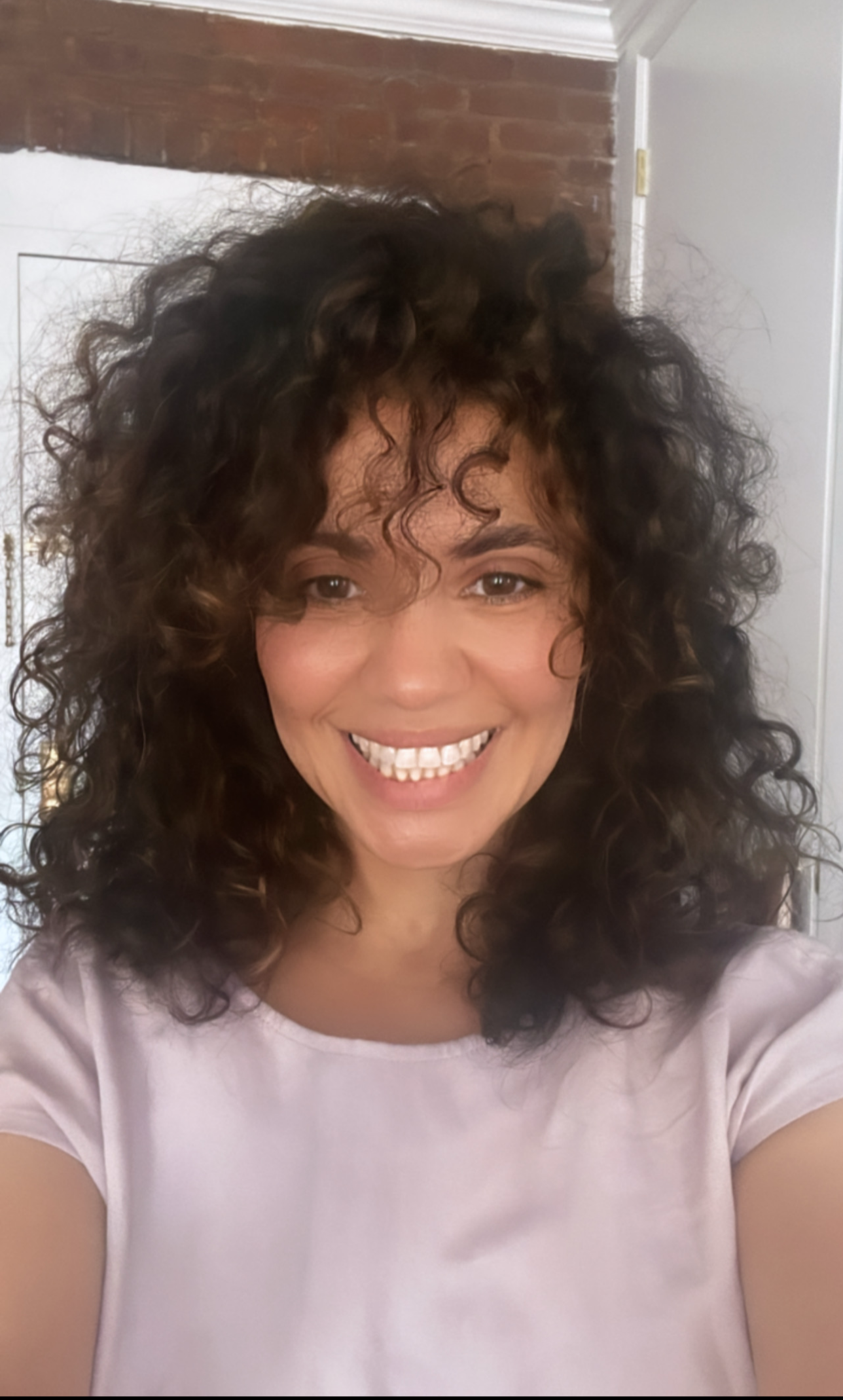 Smiling person with curly hair in a light pink shirt, standing indoors against a brick wall.
