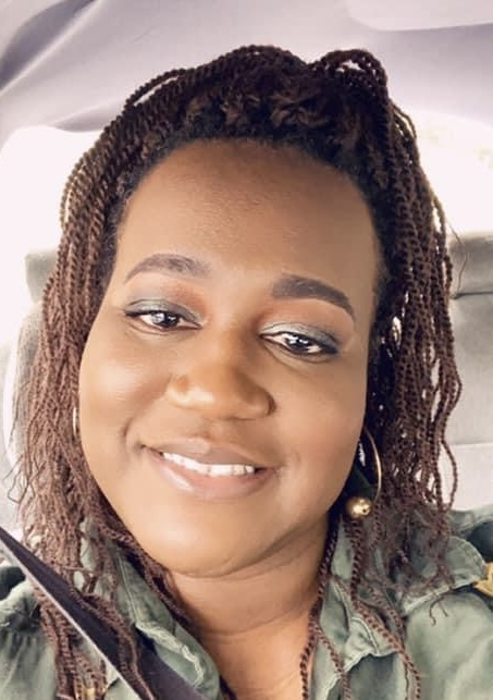 Smiling person with braided hair in a green jacket, seated inside a car.