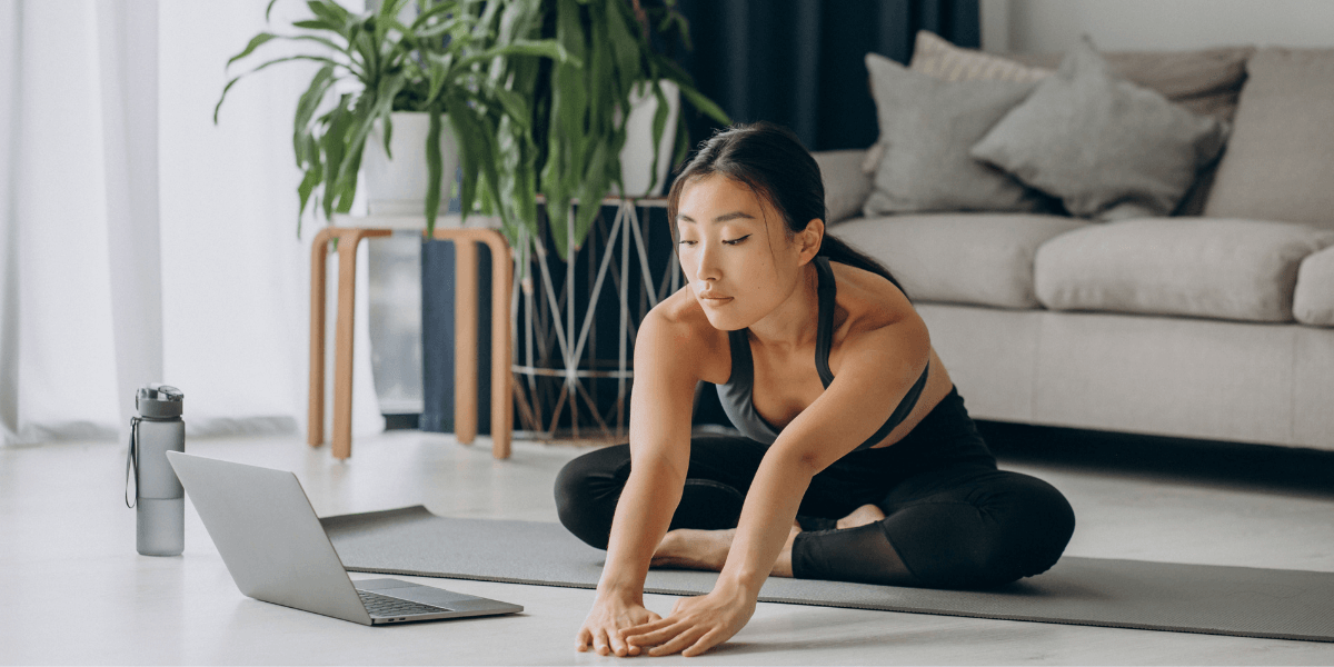 Woman stretching on a yoga mat beside a laptop in a bright living room