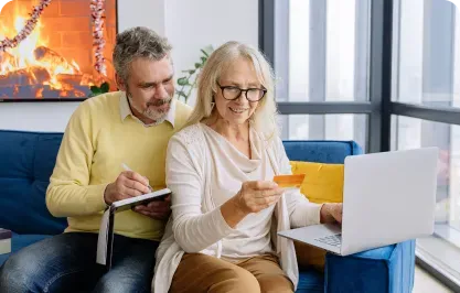 Two people on a couch using a laptop and tablet in a bright living room.