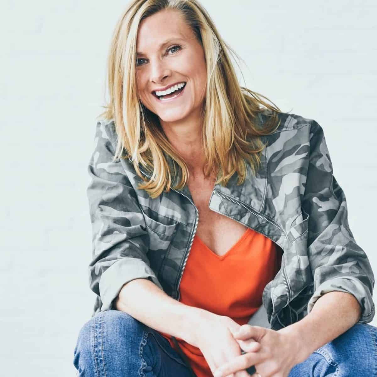 Woman with blonde hair, smiling. Wearing camouflage jacket, orange top, and blue jeans, seated against white background.