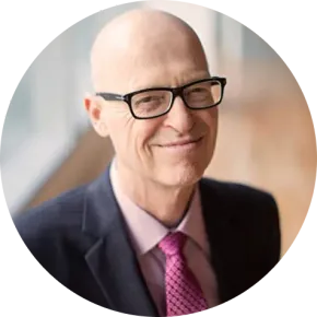 Portrait of a bald man wearing black glasses, a dark suit, and pink tie, smiling softly