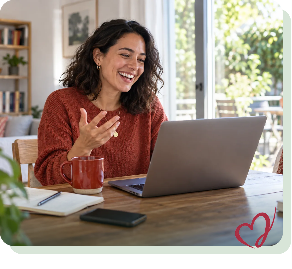 Woman smiling during a video call at a desk with a laptop, red mug, and notebook