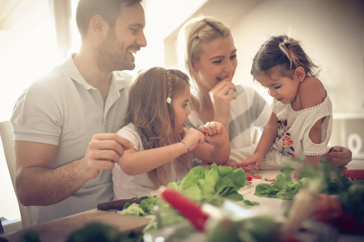 Family preparing salad together at a kitchen table, smiling and sharing vegetables.