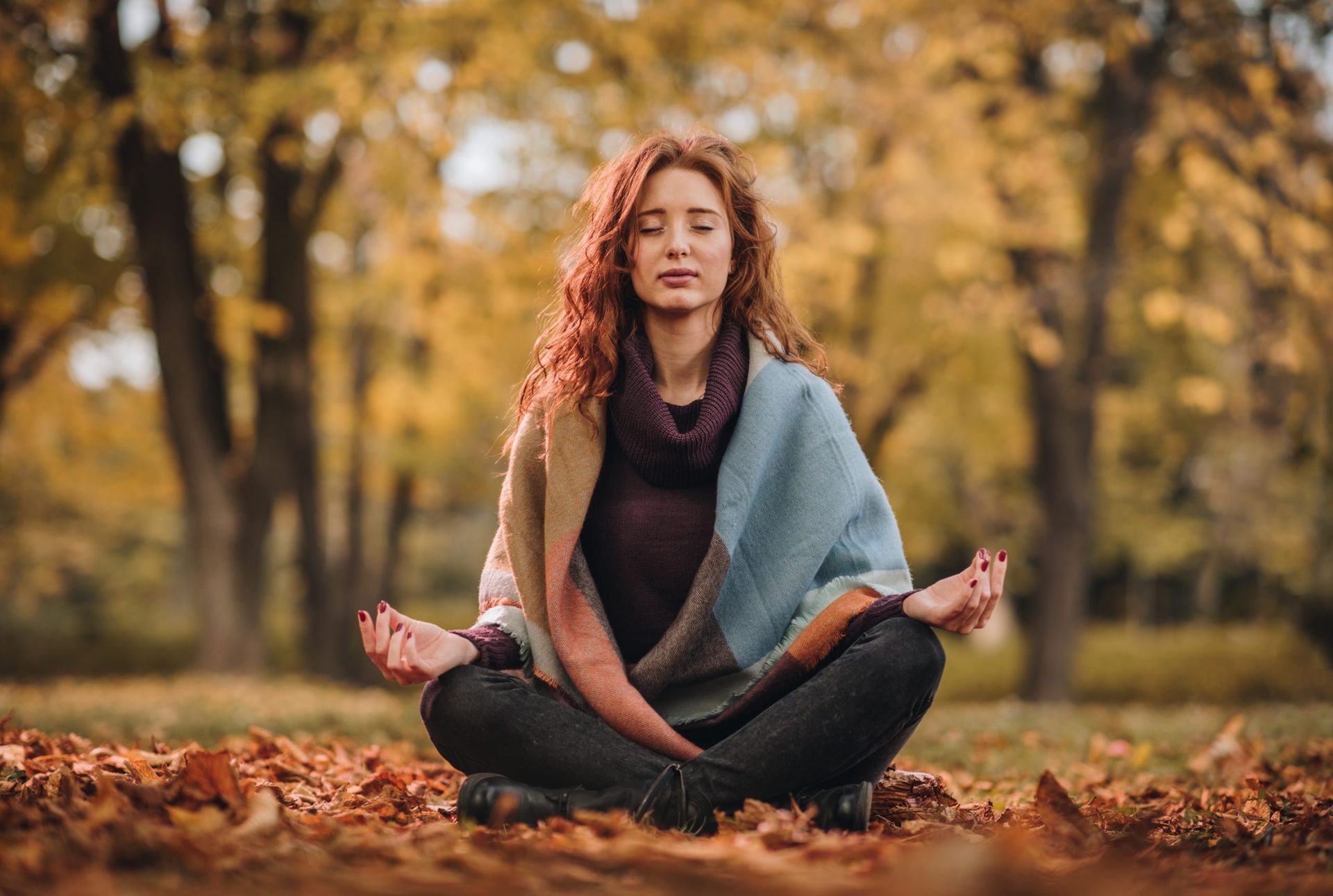 Person meditating cross-legged on autumn leaves in a park, wrapped in a blue shawl.