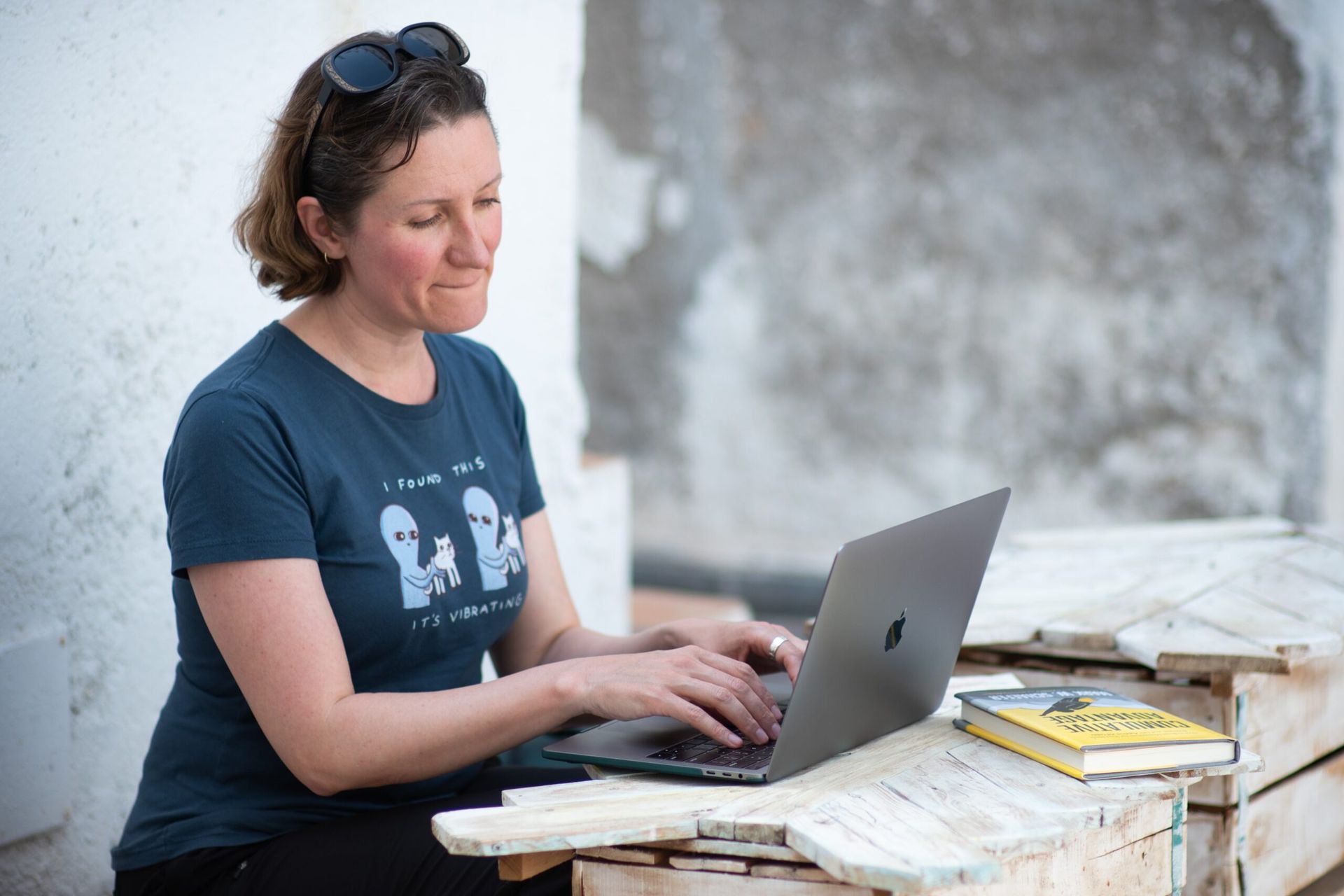 Woman using laptop outdoors, wearing blue shirt with aliens, sunglasses on head, yellow book nearby.